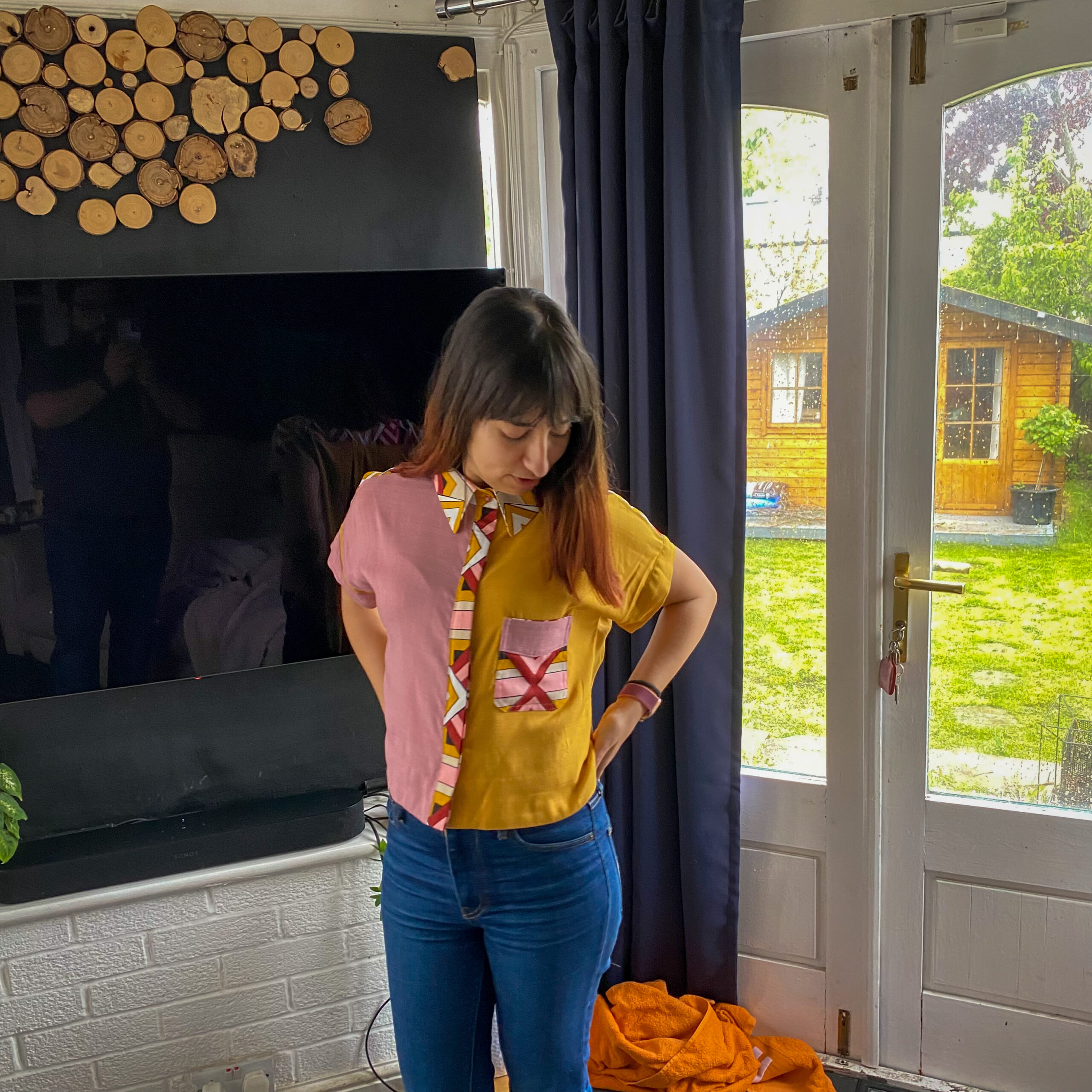 Andreia, a white woman with brown hair is looking down with hands in the back pockets of her jeans. She is wearing a half yellow and half pink shirt with african wax fabric